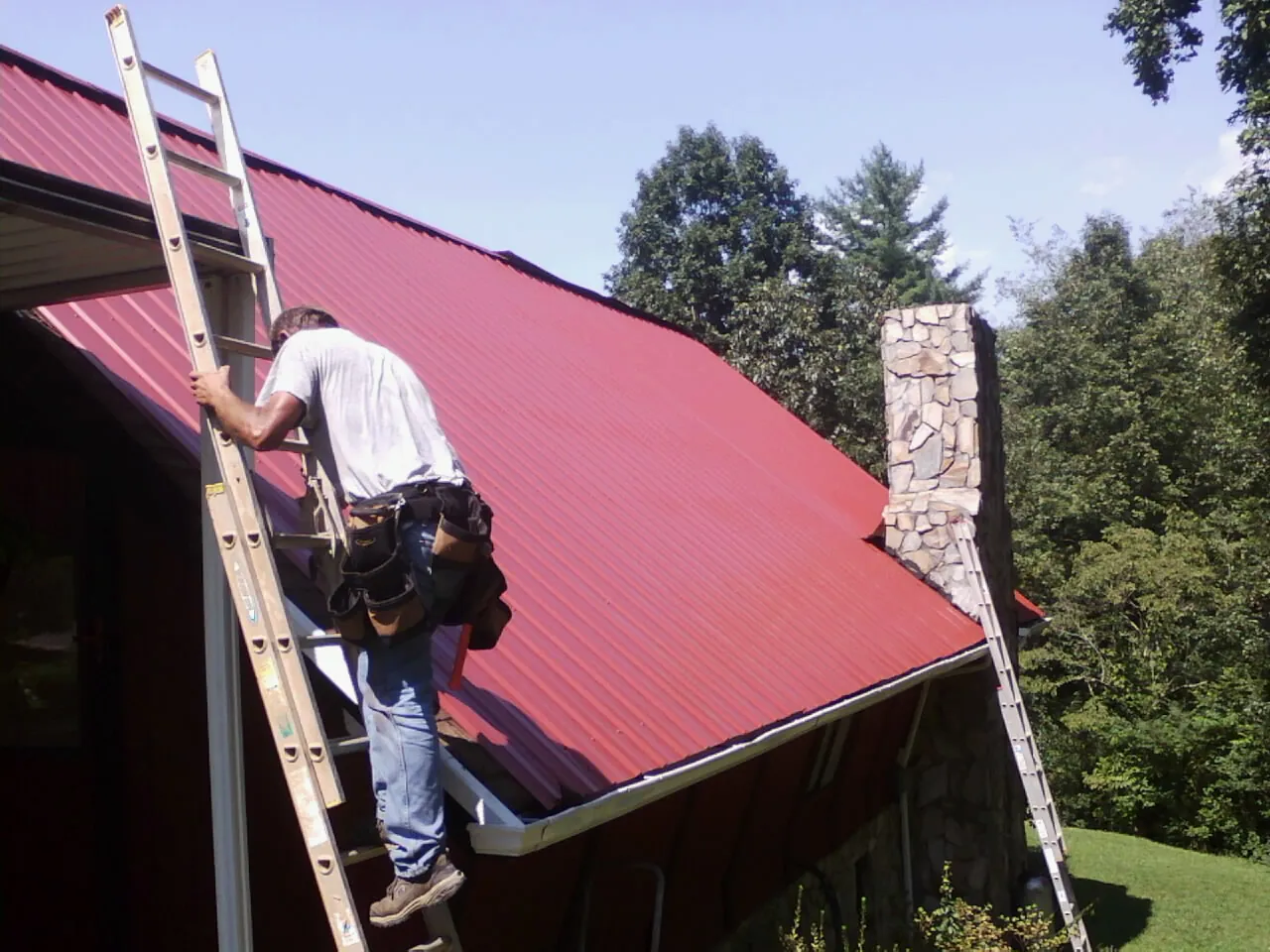 Roofer on a ladder beside a red metal roof and stone chimney — Soffit & Fascia Repair by Roofing Brothers in Saint Joseph